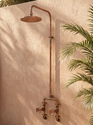 Rustic copper shower system mounted on a beige wall with a plant in the background