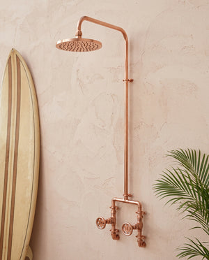 Copper shower system mounted on a beige wall with a plant in the corner