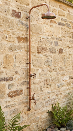 Outdoor shower system mounted on a stone wall with plants around.