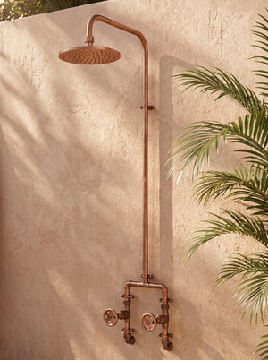 Vintage copper shower system mounted on a textured wall with greenery.