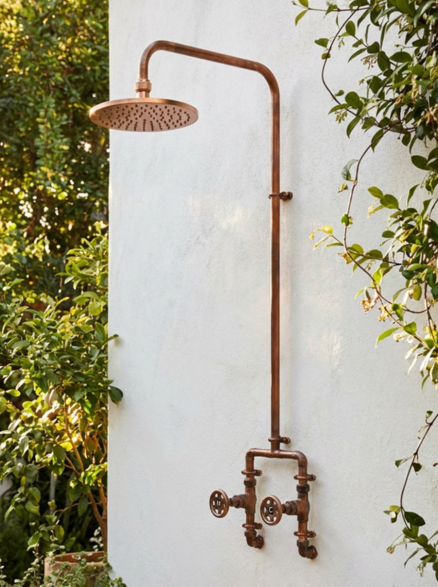 Rustic copper shower system mounted on a white wall with greenery around.