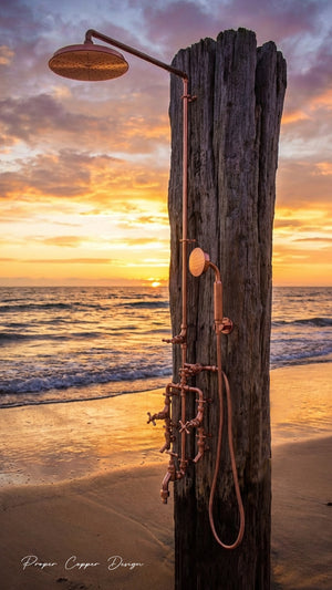 Copper shower head and fixtures attached to a wooden post on a beach at sunset.