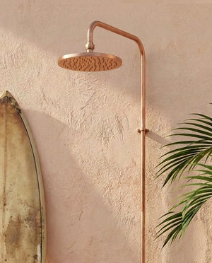 Copper shower head and arm mounted on a textured wall with a plant in the corner.