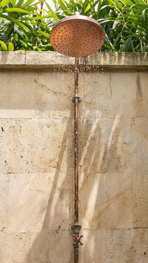 Outdoor copper shower head attached to a pipe against a stone wall with greenery in the background